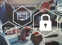 Two people are sitting at a desk, working on a computer that displays graphs and data. Above them, floating hexagonal icons represent digital security, including a lock symbol for cybersecurity, an email icon, a chat bubble, a mobile phone, and a computer screen. The image suggests they are discussing or analyzing cybersecurity, data protection, or IT security strategies.