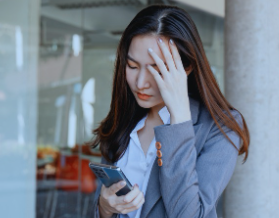 A woman wearing a gray business jacket and a white blouse looks worried or scared. She rests her left hand on her head while holding a cellphone in her right hand. Her expression suggests she has seen something on her phone that has frightened her.