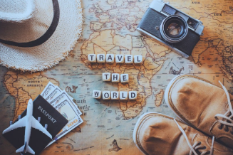 A top-down image shows a world map as the background. Placed on top are travel-related items, including a passport, hundred-dollar bills, a camera, hiking shoes, and a small toy airplane. A white woven fedora hat rests nearby, adding to the travel theme. In the center, the words "Travel The World" are clearly displayed.