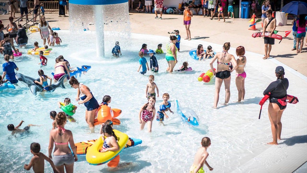 A group of children and adults play and hang out in a shallow swimming pool.