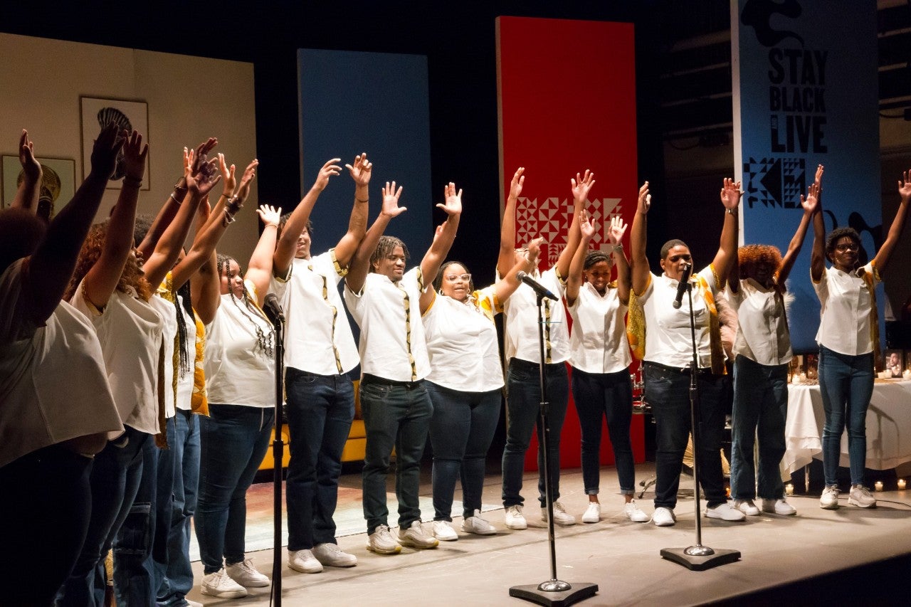 A group of Black teens in white t-shirts and jeans stand in front of microphones with theirs arms raised.