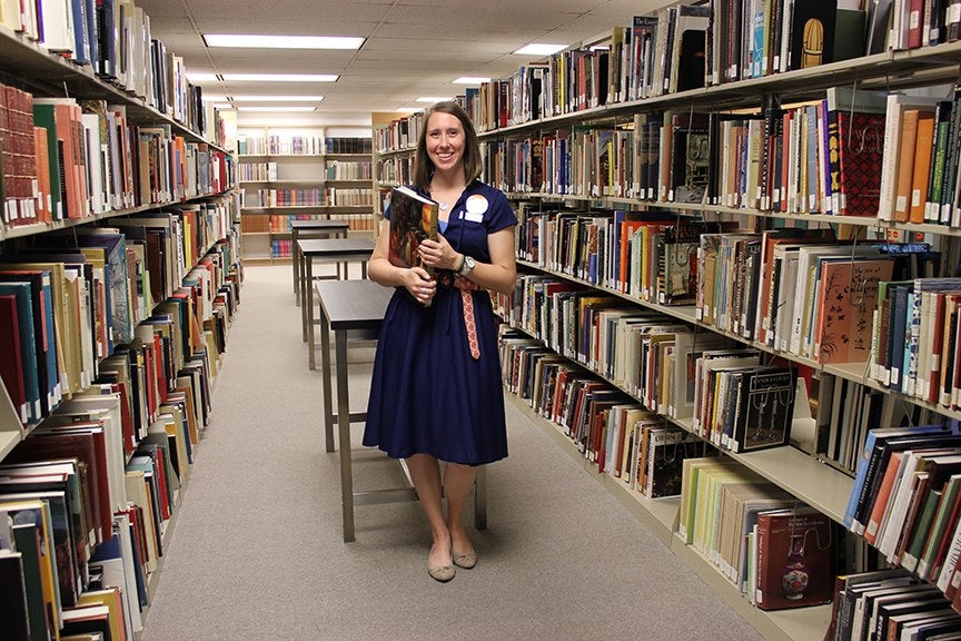 Librarian wearing a dress and holding a book standings in between two stacks of library books.