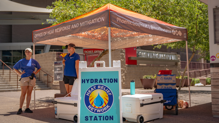 Two individuals standing under a tent with a cooler in front of them and a sign that reads "Hydration Station"