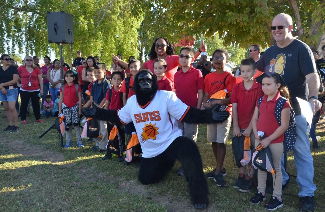  A group of young children wearing matching red t-shirts with "Downs Diamondbacks" printed on them stands outside on a grassy field, accompanied by teachers and parents. They hold a large red banner that reads "GLD Kindergarten Smarties," decorated with images of Smarties candies. A mascot, dressed as a snake wearing a black sports jersey with the number "00," stands in the center, waving. The group is smiling and posing for the camera with trees and a chain-link fence in the background.