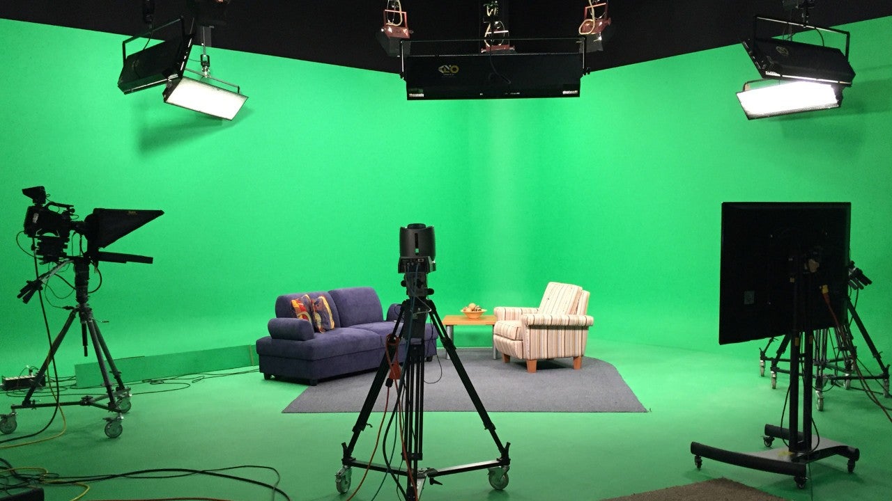 A green screen room set up with cameras, lights, and two seats facing each other.
