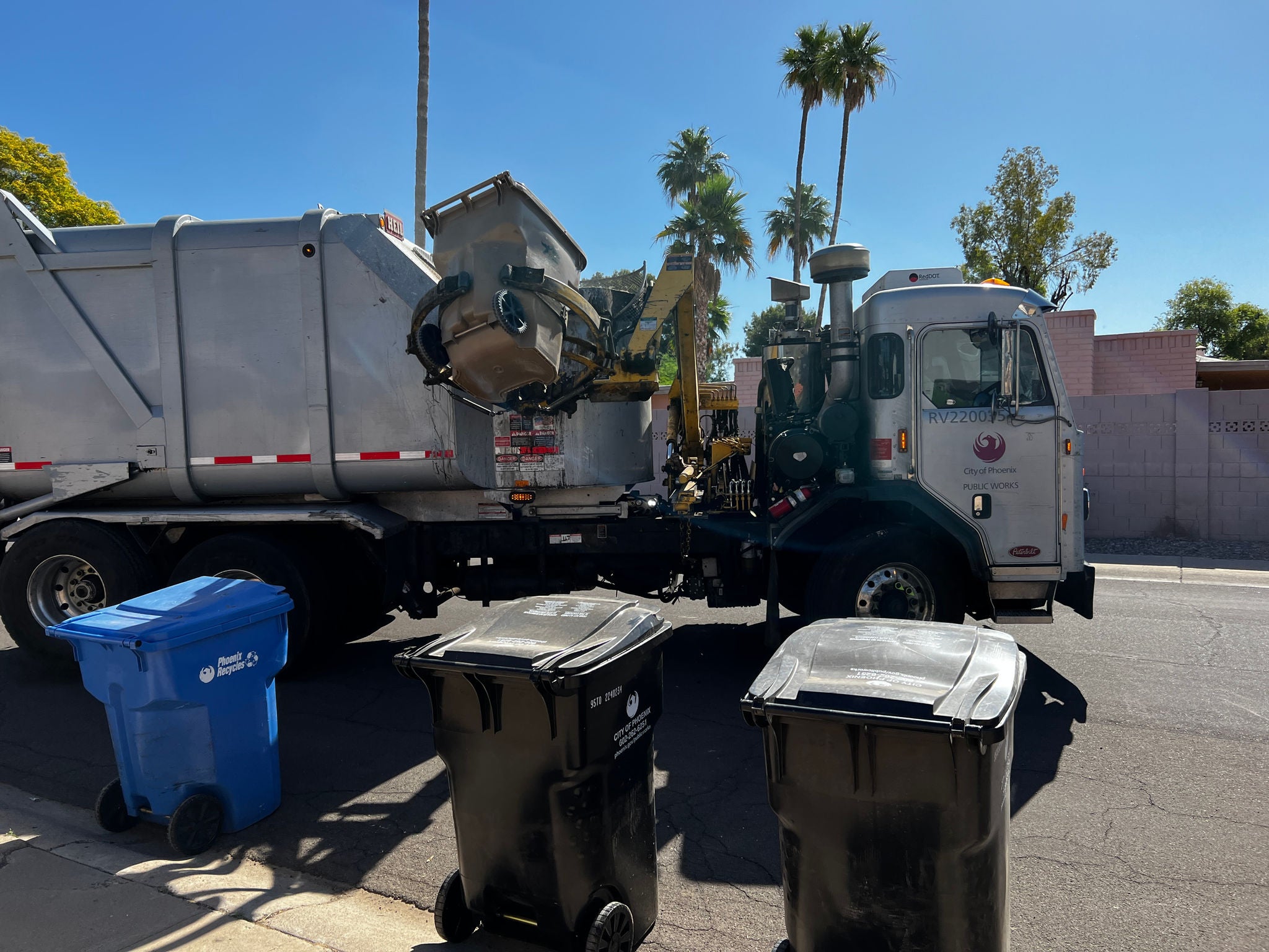A garbage truck collects a solid waste container among three standing at the curb.