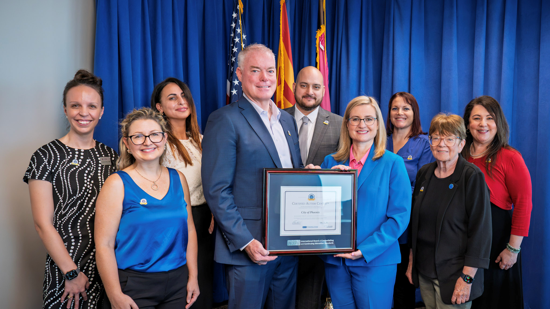 Group photo that includes Phoenix Leadership including Mayor Kate Gallego, Councilwoman Debra Stark , Deputy City Manager Dave Matthews, Equal Opportunity Department Director Judy Boros, IBCCES staff and other Phoenix staff standing in front of a blue curtain with the US and Arizona State flags. 